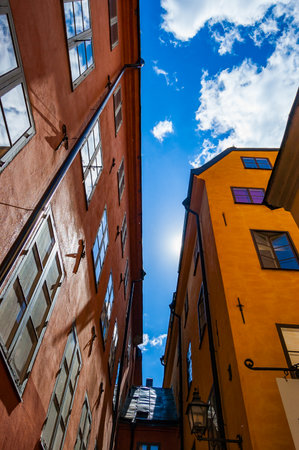 View from below on cozy narrow medieval street with connected yellow orange red buildings facades in Gamla stan, Old Town of Stockholm, Sweden with bright blue sky and sun beams on the backgroundのeditorial素材