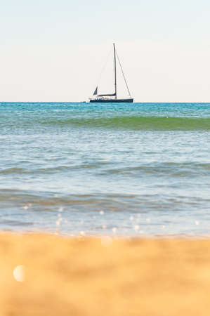The focus on the sailing yacht in the Tyrrhenian sea waters near the Porto Ercole in the Province of Grosseto, Tuscany, Italy and blurred sea waves and beach sand on the foregroundの写真素材