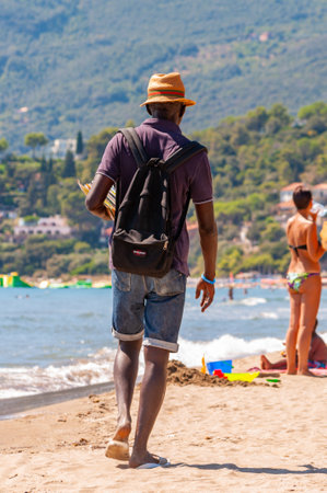 Tenda Gialla, Province of Grosseto, Tuscany, Italy - September 03, 2019: Man holding showing offering selling various printed books while walking on the beach between resting touristsのeditorial素材