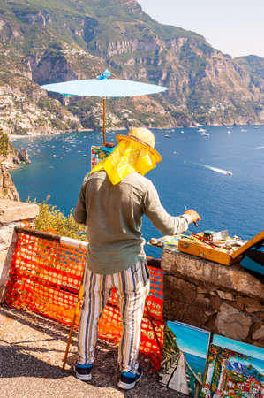 Positano, Italy - September 05, 2019: The artist painting scenery of a beautiful Positano city coast scenic rocky surroundings on one of a cliffs with blue sea waters full of yachts and high mountainsの写真素材