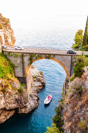 Positano, Italy - September 05, 2019: View on Fiordo di Furore arc bridge built between high rocky cliffs above the Tyrrhenian sea bay in Campania region. Cars driving on bridge, boat floating underの写真素材