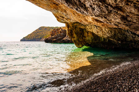 Amazing seascape view from unique sea cave overhanging above water on famous Cala Bianca beach scenic surroundings. Crystal clear sea water waves washing pebbles inside the massive rocky cave. Italyの写真素材