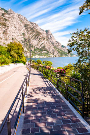 Cozy city street of Limone Sul Garda with paving stone sidewalk, blooming flowers on a metal railings, growing trees with amazing Garda lake and dolomite mountains on the backgroundの写真素材