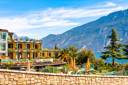 Limone Sul Garda, Lombardy, Italy - September 12, 2019: Cozy city street of Limone Sul Garda with growing trees, comfortable hotels with the amazing Garda lake and dolomite mountains on the backgroundの写真素材