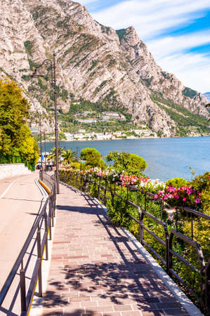 Cozy city street of Limone Sul Garda with paving stone sidewalk, blooming flowers on a metal railings, growing trees with amazing Garda lake and dolomite mountains on the backgroundの写真素材