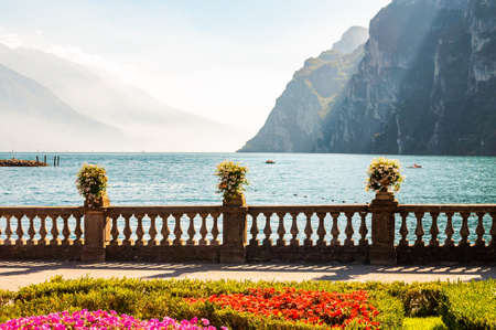 Garda lake promenade with colorful flowerbeds with growing and blooming plants, classic stone fence built on the edge with flowerpots with blooming flowers. Garda lake and high mountains on backgroundの写真素材