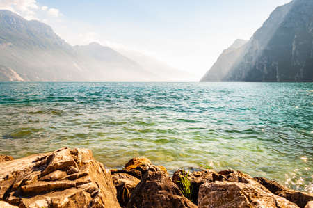 Rocky stones lying on the shore of beautiful Garda lake in Lombardy, Italy surrounded by high dolomite mountains. Sun rays penetrating from above the rocks and warming misty fog above the waterの写真素材