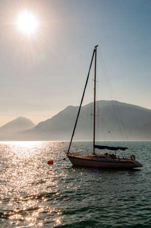 Sailing yacht floating near shore of misty Garda lake with high dolomite mountains with sun shining...