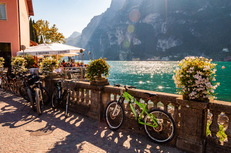 Riva del Garda, Lombardy, Italy - September 12, 2019: Bicycles parked near the stone fence railings near the outdoor restaurant full of people on Garda lake promenade with high mountains on backgroundのeditorial素材