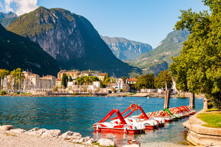Riva del Garda, Italy - September 12, 2019: Beautiful Riva del Garda cityscape with vibrant red pedal boats parked in a row on the beach and city surrounded by high dolomite mountains on backgroundのeditorial素材