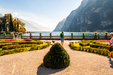 Riva del Garda, Lombardy, Italy - September 12, 2019: People walking by Garda lake promenade full of cozy alleys with growing and blooming plants, classic stone fence with flowerpots built on the edgeのeditorial素材