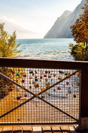 Riva del Garda, Lombardy, Italy - September 12, 2019: Love and marriage locks hanging on metal grid on wooden railings in Riva del Garda city with beautiful lake waterscape on the backgroundのeditorial素材