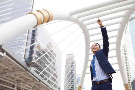Businessman smiling and raising his fist with number 1 in the air, with office building background - business success, achievement, and win conceptsの写真素材
