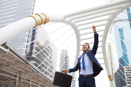 Businessman smiling and raising his fist with number 1 in the air, with office building background - business success, achievement, and win conceptsの写真素材