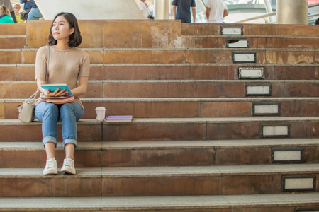 Beautiful Asian female college student holding her books and a cup of coffe sitting at stairs, people education learning high school program smart teenager conceptの写真素材
