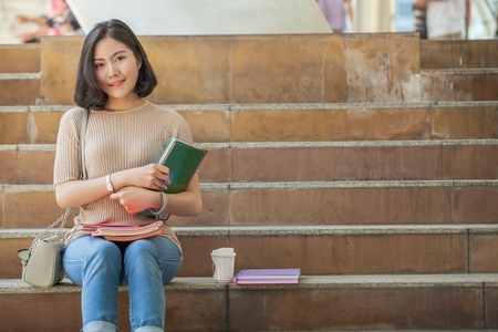 Beautiful Asian female college student holding her books and a cup of coffe sitting at stairs, people education learning high school program smart teenager conceptの写真素材