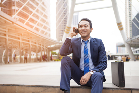 Businessman sitting on stair during callling the phone, with office building background - business success, achievement, and win conceptsの写真素材
