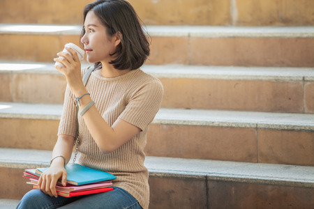 Beautiful Asian female college student holding her books and a cup of coffe sitting at stairs, people education learning high school program smart teenager conceptの写真素材