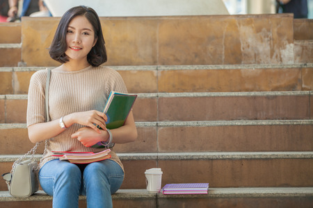 Beautiful Asian female college student holding her books and a cup of coffe sitting at stairs, people education learning high school program smart teenager conceptの写真素材