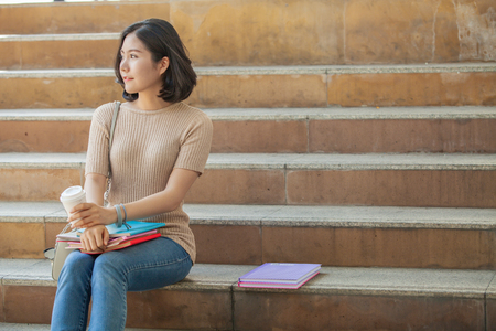 Beautiful Asian female college student holding her books and a cup of coffe sitting at stairs, people education learning high school program smart teenager conceptの写真素材