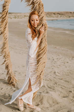Young asian girl in a white wedding dress in a boho style, in the desert. With red long hair, and makeup. Boho style wedding arch. Sea wedding concept.の写真素材