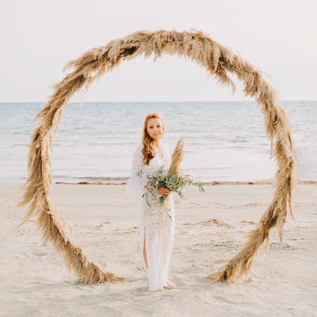 Young asian girl in a white wedding dress in boho style, on the beach. With red long hair, and makeup. Boho style wedding arch. Sea wedding concept.の写真素材