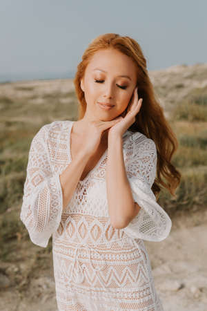 Young asian girl in a white wedding dress in a boho style, in the desert. With red long hair. With beautiful hairstyle and makeup. Makeup with nice false eyelashes. Sea wedding concept.の写真素材
