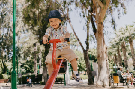 Child boy ride on a swing in the park. Wearing a hat, shirt and shorts. In the summer at the playground. Summer, vacation, outdoor recreation.の写真素材