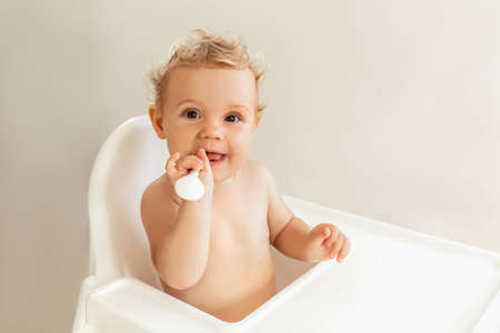 Happy baby girl sitting in a high chair and holding a spoon in her mouth. Babys first food. 7 months...