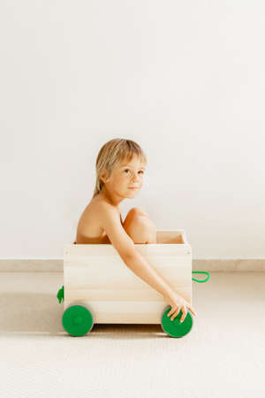 Blonde baby boy in a wooden box, with green wheels for toys, on a white background.