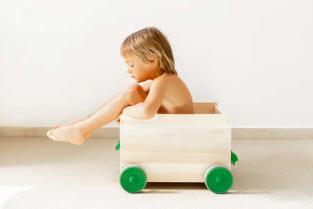 Blonde baby boy in a wooden box, with green wheels for toys, on a white background. Sad and offended childの写真素材