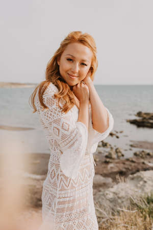 Young asian girl in a white wedding dress in a boho style, in the desert. With red long hair. With beautiful hairstyle and makeup. Makeup with nice false eyelashes. Sea wedding concept.の写真素材