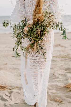 Bride in white boho style dress with a bouquet of artichoke and bamboo on the beach. Sea wedding concept.の写真素材