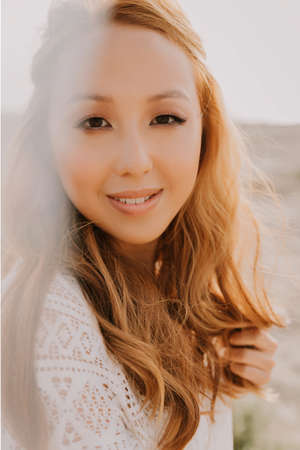 Young asian girl in a white wedding dress in a boho style, in the desert. With red long hair. With beautiful hairstyle and makeup. Makeup with nice false eyelashes. Sea wedding concept.の写真素材