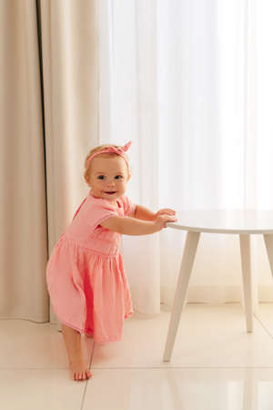 Child girl in a pink dress stands holding the table. 8 months old baby learns to walk and stand at the support.の写真素材