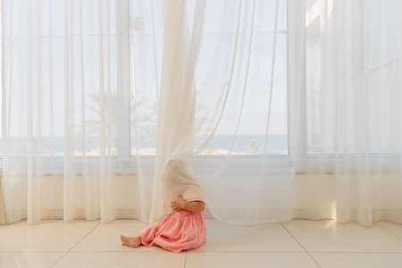Happy baby girl in a pink dress with white curly hair playing hide and seek with curtains. 8 months baby.の写真素材