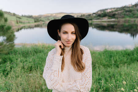 Young adorable girl in white dress and black hat, in spring, posing for camera. female portrait outdoor in mountain lake. Green fresh grass. Young unusual person enjoying natureの写真素材