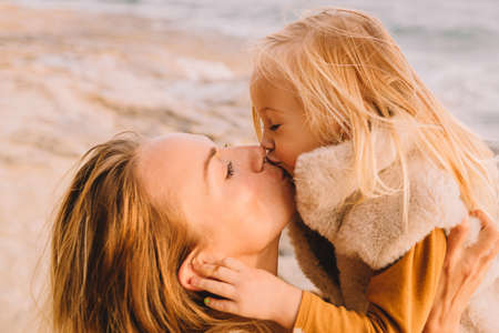 Mother with baby daughter next to the sea smiling. Baby girl with blonde hair, in a warm fur vest. Spring time, sun is shining. Travel abroad. Happy young family.の写真素材