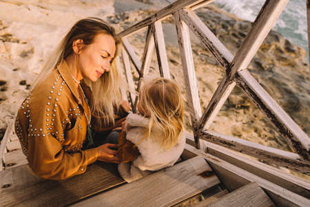 Mother with baby daughter next to the sea smiling. Baby girl with blonde hair, in a warm fur vest....