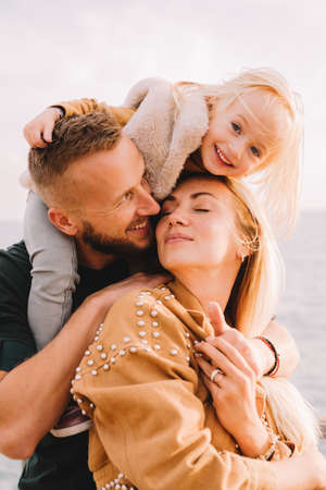 Mother, dad and baby daughter next to the sea smiling. Baby girl with blonde hair, in a warm fur vest. Spring time, sun is shining. Travel abroad. Happy young family.の写真素材