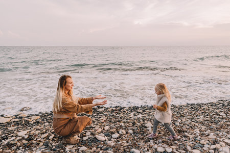 Mother with baby daughter next to the sea smiling. Baby girl with blonde hair, in a warm fur vest. Spring time, sun is shining. Travel abroad. Happy young family.の写真素材