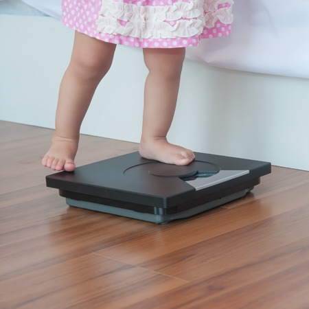 Cute child 1 year and 2 months step up on personal Weight Scale in bedroom nexto white bed with her mother.の写真素材