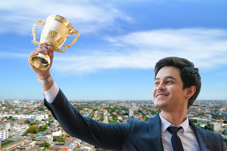 Success business man holding golden trophy cup with right hand, Business executives rejoice in their leadership in business competition.  Congratulations to the gold trophy with blur city town.の写真素材