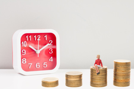 Selective focus miniature woman sitting on the golden coins stack and blur red alarm clock, business conceptual money growth up increases with time.の写真素材
