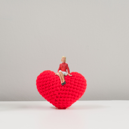 Selective focus miniature girl woman wearing long red cardigan sitting on top of knitting wool red heart, on white table and grey background.の写真素材