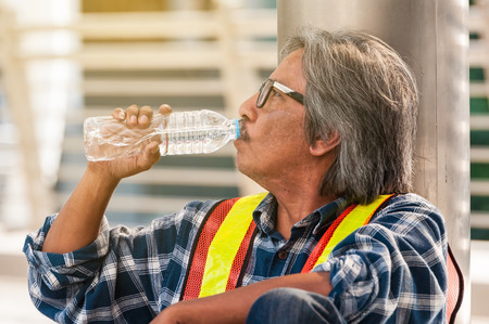 Middle Asian engineer has dark grey hair sitting and raise pure water from plastic bottle to thirsty in middle construction areaの写真素材