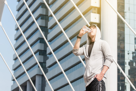 Young Asian man wearing grey sport jacket with hood drinking water from plastic bottle with right...