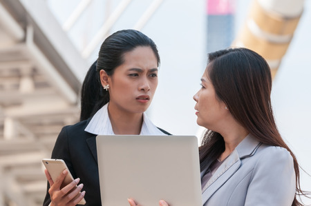 Unhappy young and middle Asian businesswomen have crisis problem after check their work email from smartphone and laptop in middle city town.の写真素材