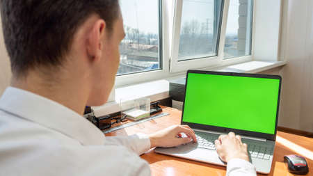 A young man in a white shirt working in an office using a laptop computer with a green screen on the...