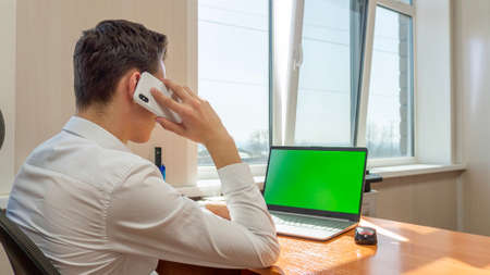 A young man sitting in office talking on the phone in front of a computer with a green screen. High quality photoの写真素材